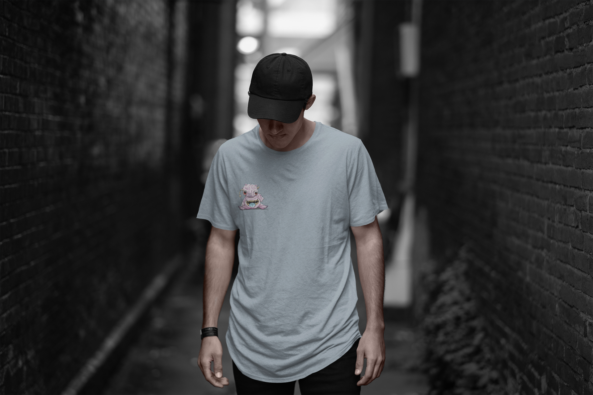 A man in a cap standing in a desaturated alley in a pale blue shirt. With a pink bublegum looking alien creature on the front of the shirt small "pocket sized" display on the front left